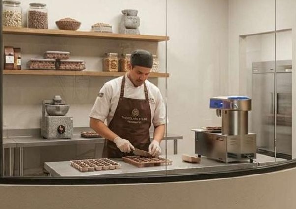 Chocolatier working at the glass-walled chocolate atelier with a tempering machine and shelved ingredients